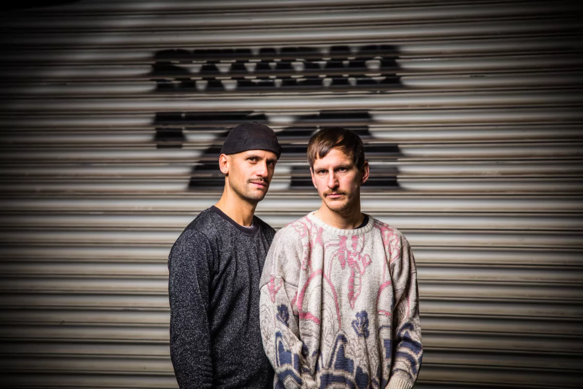Das Bild zeigt links Daniel Ernesto Mueller und rechts Simon Hartmann, die vor einem beigen Rolltor stehen mit einem schwarzen Graffitti im Hintergrund.