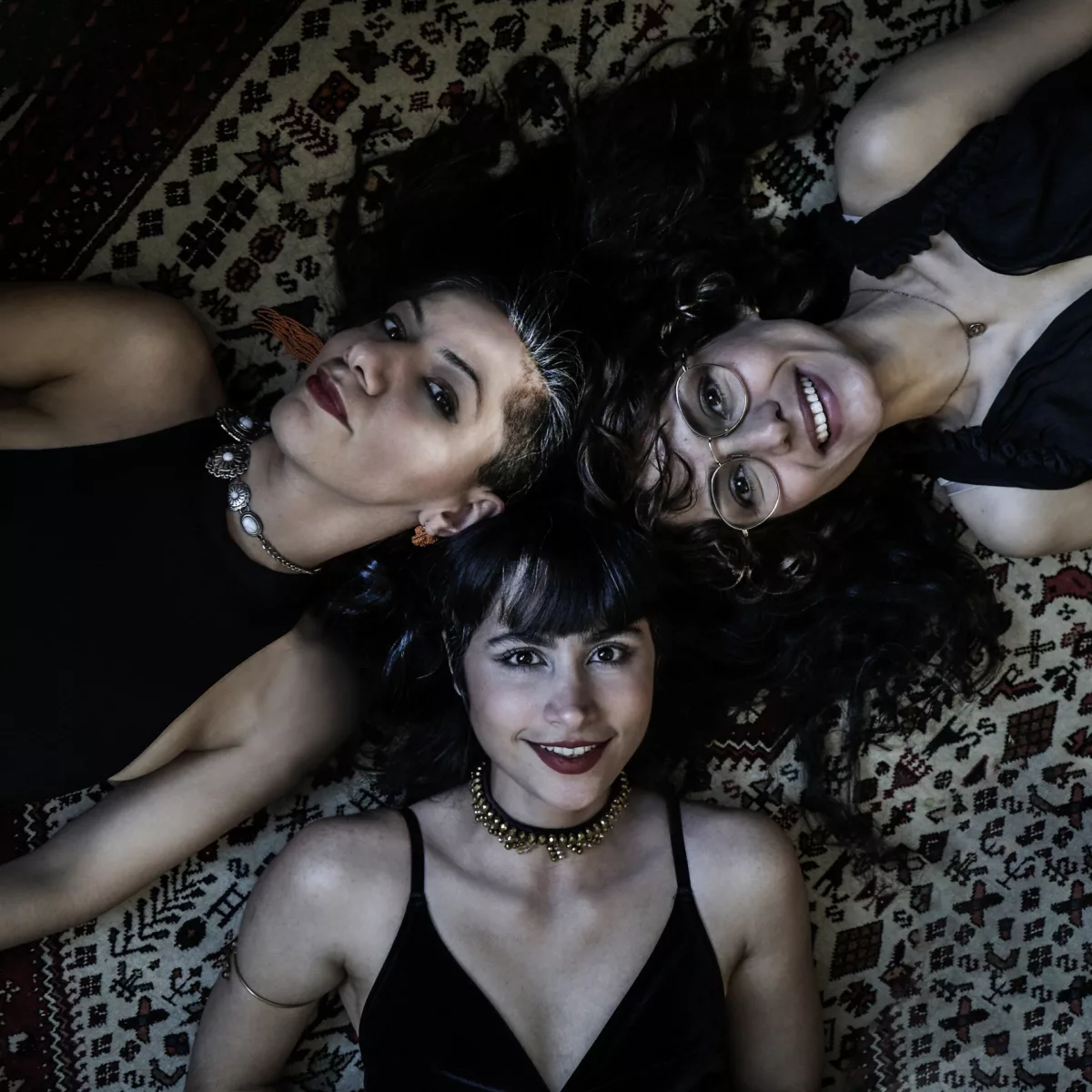Das Bild zeigt drei junge Frauen, die auf dem Rücken liegen und in die Kamera schauen. Alle drei haben langes dunkles Haar und tragen schwarze ärmellose Oberteile. Sie liegen auf einem gemusterten Teppich.