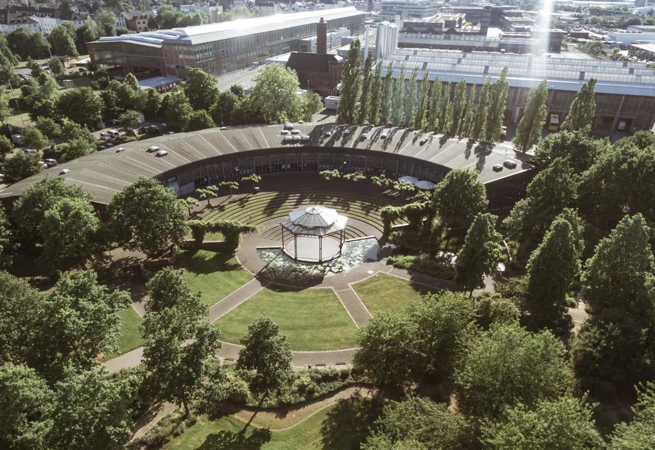 Das Foto zeigt den Ringlokschuppen Ruhr aus der Vogelperspektive, sodass die Halbkreis-Form des Gebäudes sichtbar wird. Das Gebäude ist umgeben von einem Park mit Bäumen.