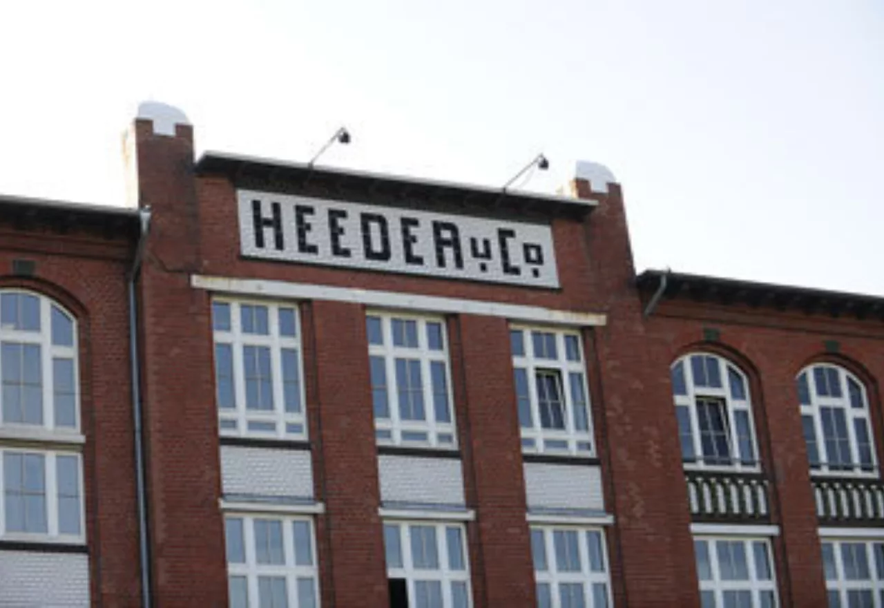 Das Foto zeigt die Außenansicht der Fabrik Heeder. Das Gebäude besteht aus rotem Backstein mit vielen Fenstern. Auf weißem Hintergrund ist aus Backsteinen der Schriftzug "HEEDER u CO" zu lesen.