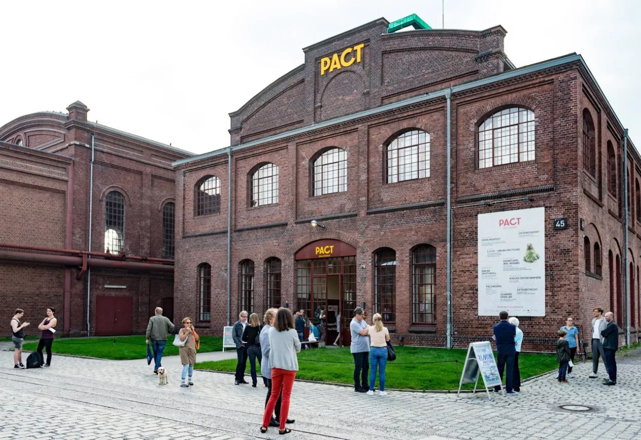 Das Bild das Gebäude von PACT Zollverein. Die alte Waschkaue aus rot-braunen Backsteinen. Auf dem Platz davor stehen mehrere Personen.
